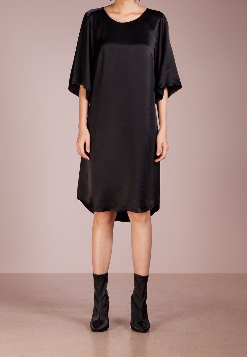Femme portant une robe ample en satin noir longueur genou avec de larges manches et des bottines noires assorties sur un fond uni.