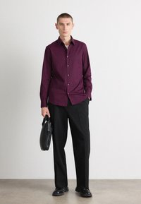 Burgunderfarget skjorte med lange ermer, kombinert med svarte bukser og svarte sko; modellen holder en svart lærveske.