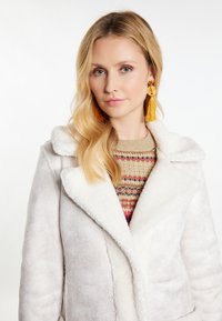 Giacca in finta pelliccia bianca con un ampio colletto, indossata sopra un maglione decorato con tonalità terrose e accenti rossi.