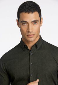 Camisa de hombre verde oscuro de botonadura con una tela texturizada, cuello y botones negros, que presenta un diseño ajustado y detalles de patrón sutiles.