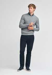 Grå V-ringad tröja i stickad tyg, kombinerad med mörka byxor och avslappnade skor. Slät textur och figursydd design med subtil logotypdetalj.