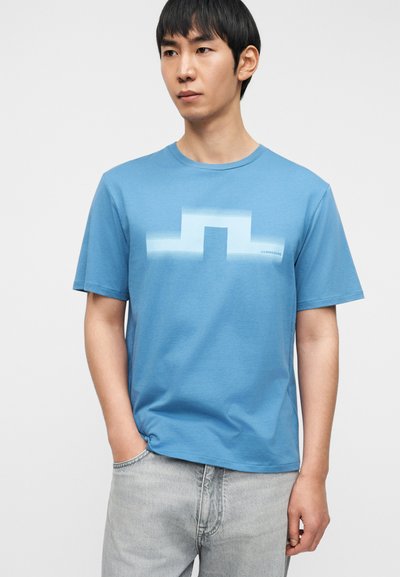 Γαλάζιο βαμβακερό T-shirt με γεωμετρικό σχέδιο στο κέντρο. Κοντά μανίκια και λαιμόκοψη τύπου crew. Φορεμένο με ανοιχτό γκρι τζιν.