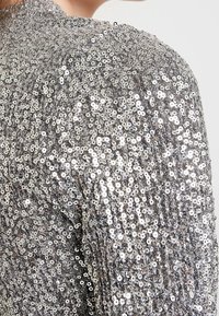 Silberner, paillettenbesetzter Stoff mit strukturierten Oberflächen bedeckt den Schulterbereich und zeigt runde Pailletten, die das Licht in unterschiedlichen Richtungen reflektieren.