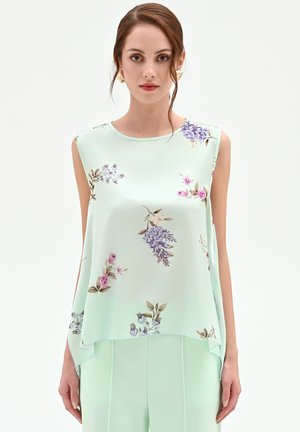 Femme portant un haut sans manches vert clair avec des imprimés floraux roses et violets, assorti à un pantalon vert clair, sur un fond blanc.