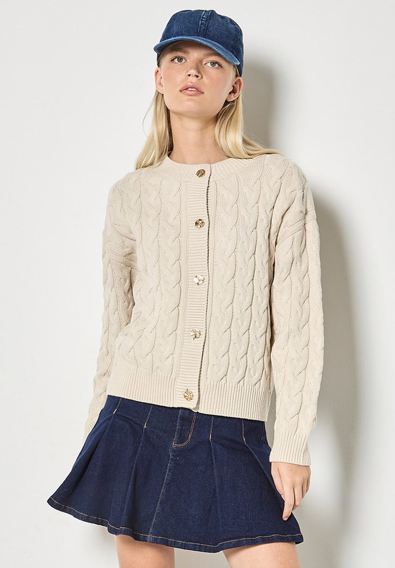 Beige knitted cardigan with cable pattern, round neckline, and five gold-tone buttons. Paired with a blue denim skirt and cap.