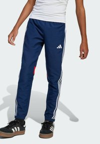 Marina byxor i träningsdräkt med vita sidostreck, med röd detalj och en Adidas-logotyp på vänstra låret. Elastiska mudd vid anklarna.