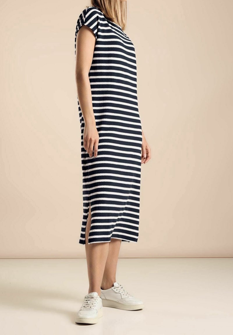 Femme portant une robe midi rayée noire et blanche avec des fentes latérales et des baskets blanches, debout sur un fond beige.