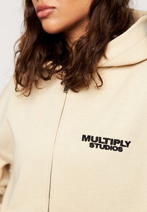 Frau mit schulterlangem, lockigem braunem Haar, die einen beigen Kapuzenpullover mit Reißverschluss trägt, auf dessen Brust "MULTIPLY STUDIOS" in Schwarz aufgestickt ist.