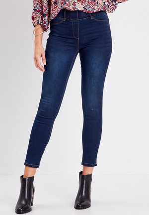Jean skinny bleu foncé à taille haute, avec un style cinq poches et des coutures contrastées. Assorti à des bottes noires à la cheville.