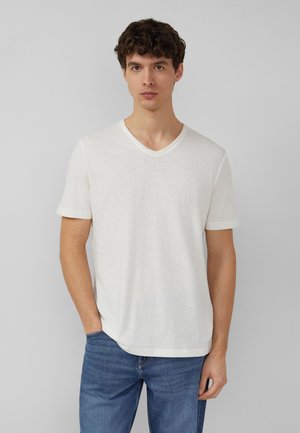 Junger Mann mit kurzen lockigen Haaren, der ein schlichtes weißes V-Ausschnitt-T-Shirt und blaue Jeans trägt, steht vor einem neutralen grauen Hintergrund.