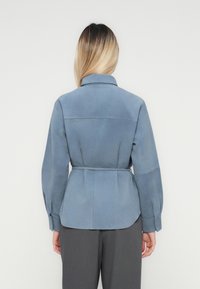 Vrouw met blond ombre haar staat met haar rug naar de kijker, gekleed in een lichtblauwe jas met ceintuur en donkergrijze broek, tegen een plain witte achtergrond.
