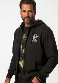 Felpa nera con zip e tasca frontale, con la scritta "HELL 80 RIDER" in lettere bianche sul lato sinistro del petto. Indossata sopra una maglietta grafica.