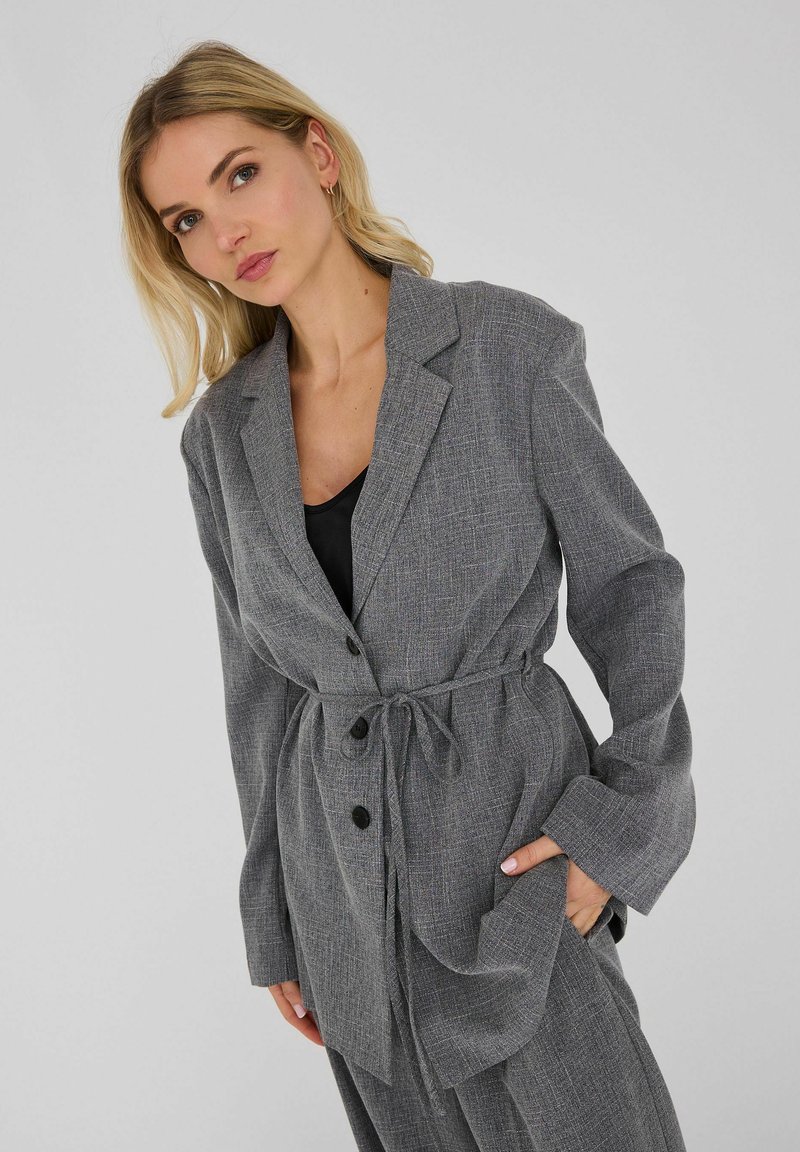 Femme blonde portant un blazer gris ceinturé et un pantalon assorti, avec un top noir en dessous, posant devant un fond uni.