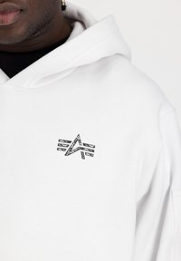 Haut à capuche blanc avec un logo graphique noir sur la poitrine. Le tissu semble doux et a un design classique avec un capuchon à cordon.