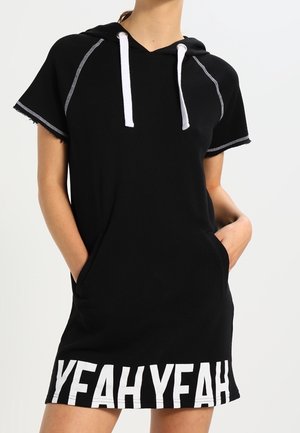 Mujer con un vestido con capucha negro de manga corta y cordones blancos, con el texto "YEAH YEAH" en el dobladillo, con las manos en los bolsillos delanteros.