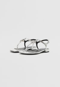Sølv flate sandaler med metallisk finish, T-stropper og dekorativt tilbehør. Svart innersåler og et minimalistisk uttrykk med en liten hæl.