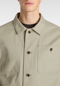 Beige cotton jacket with a pointed collar, two flap pockets, and brass buttons. The fabric has a smooth, durable texture.