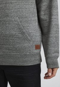 Sudadera gris con un tejido texturizado, que cuenta con un bolsillo frontal, puños acanalados y un parche de logo de cuero marrón en el dobladillo inferior.