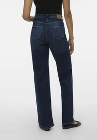 Höga, mörkblå denimjeans med rak passform, som har två bakfickor och en beige läderlappar på midjan.