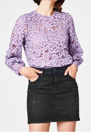 Top lungo a maniche con pizzo lilla e motivo floreale, abbinato a una gonna nera in denim aderente con dettagli sfilacciati e tasche frontali.