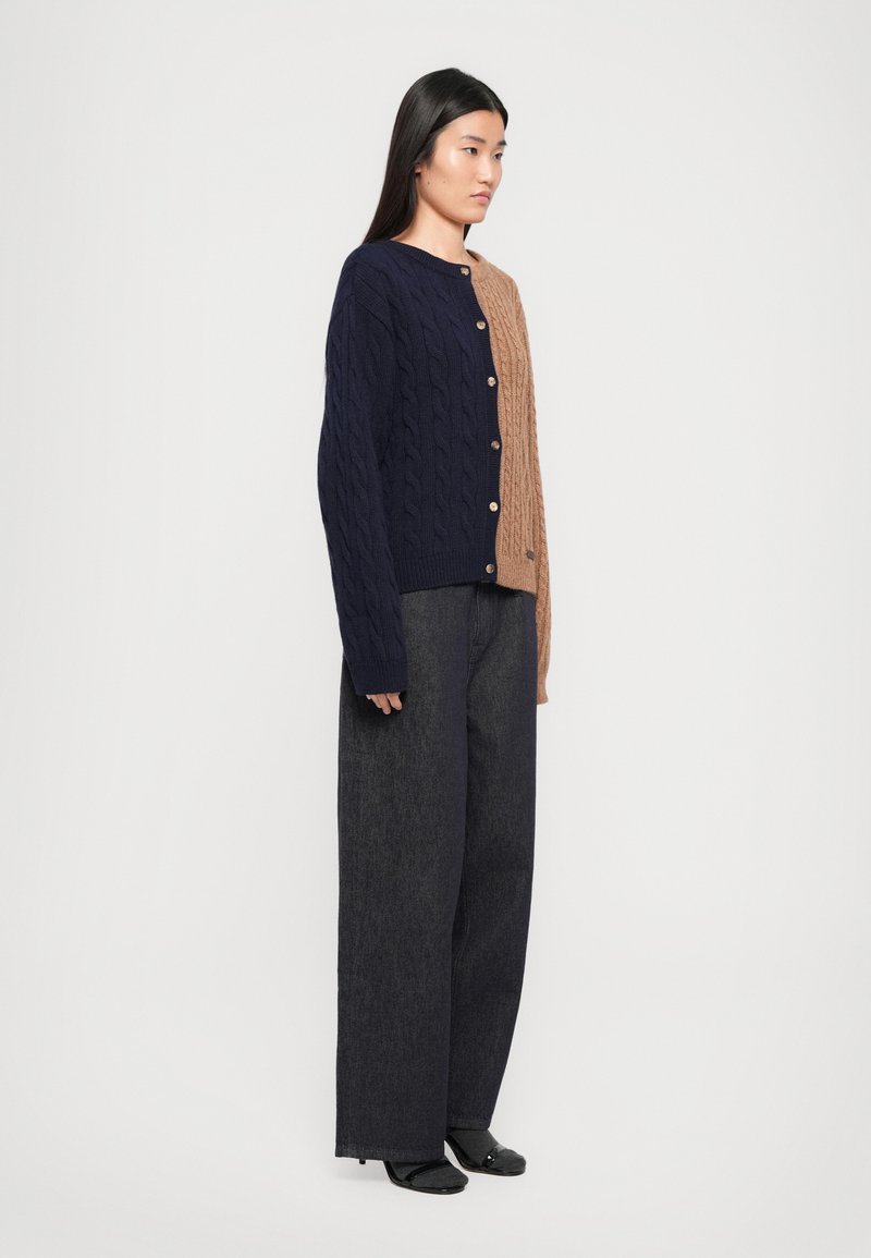 Cardigan lavorato a maglia a trecce con metà blu navy e marrone. Presenta un collo rotondo, bottoni frontali e orlo a coste. Abbinato a pantaloni larghi scuri.