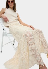 Kantrok in crème, met bloemmotieven, gelaagd ontwerp en een vloeiende silhouet. Vergezeld van een textuurloze beige mouwloze top.