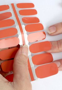 Ett set med självhäftande nagelremsor i livlig korallfärg, med blank yta och rundade kanter. En hand som håller i en remsa för applicering.