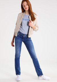 Beige bomberjack over een grijze t-shirt, gecombineerd met blauwe jeans en witte sneakers. Eenvoudig ontwerp, zachte stof en een getailleerde pasvorm.