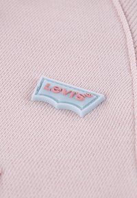 Το λογότυπο της Levi's με ανοιχτό γαλάζιο φόντο και ανάγλυφα ροζ γράμματα σε υφασμάτινη ροζ υφή.