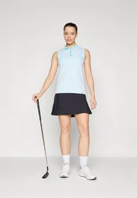 Ανοιχτό μπλε αμάνικο πόλο με υφασμάτινο μοτίβο, συνδυασμένο με μαύρη φούστα γκολφ με άνοιγμα. Το μοντέλο κρατά ένα μπαστούνι γκολφ, φοράει λευκά παπούτσια.