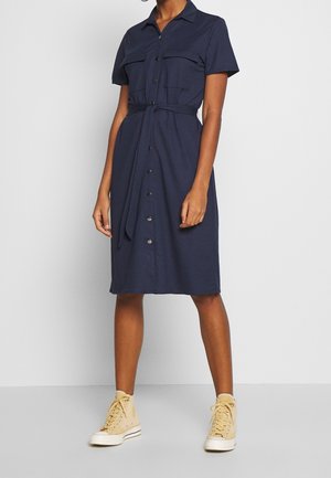 Kobieta ubrana w granatową sukienkę koszulową na guziki z krótkimi rękawami i paskiem w talii, w połączeniu z beżowymi wysokimi trampkami.