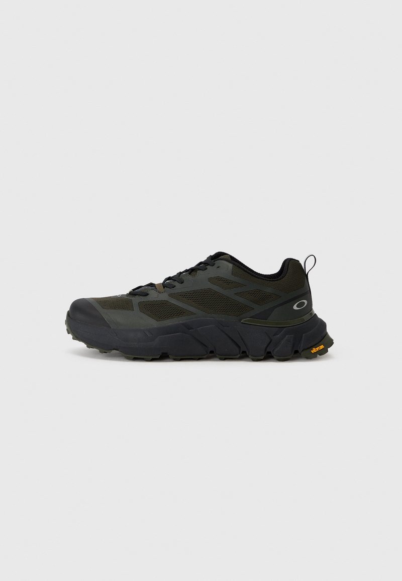 Chaussure de randonnée basse vert foncé et noire avec une semelle Vibram épaisse et une languette de tirage au talon, présentée de côté sur un fond gris uni.
