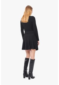 Schwarzes enges Langarmshirt kombiniert mit einem gefältelten schwarzen Rock, ergänzt durch kniehohe schwarze High Heels Stiefel. Glatter Stoff, minimalistisches Design.