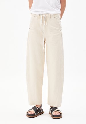 Persoon draagt wijde beige broek met trekkoord in de taille en zwarte sandalen met witte bontvoering, staande tegen een witte achtergrond.