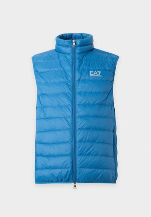 Jeune homme portant une veste matelassée bleue, une chemise blanche à manches longues, un pantalon beige, des baskets blanches et une casquette beige avec le logo EA7, debout, une main dans la poche.