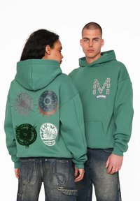 Grøn bomulds hoodie med en frontlomme, store grafiske tryk på ryggen og et stiliseret logo på forsiden.