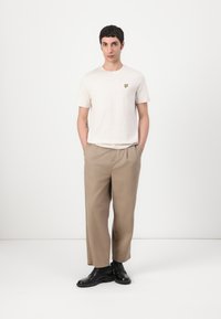 T-shirt gris clair à col rond avec un logo de papillon jaune, associé à un pantalon beige ample et des chaussures noires, se tenant devant un fond blanc.