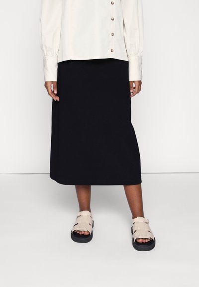 Camisa creme de botões com mangas bufantes, combinada com uma saia midi preta. Sandálias de tiras em tom claro com sola preta, mostrando os dedos dos pés.