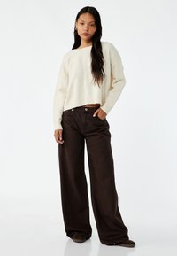 Lichtbeige gebreide trui met een cropped ontwerp en wijde mouwen. Combinatie met donkerbruine wijde broek met frontzakken en een knoopsluiting.