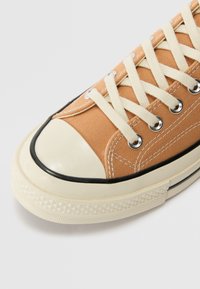 Beige kanvas sneakers med kremfarget gummitåhette og svart kant. Har hvite snøringer og metalløyer, med en strukturert såle.