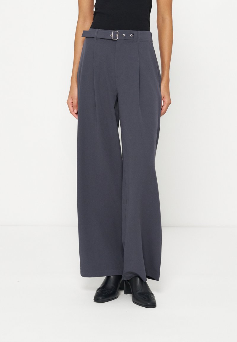 Femme portant un pantalon large gris foncé avec ceinture et des bottines noires à talons sur fond blanc.