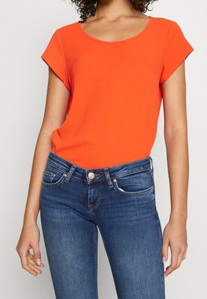 Femme portant un haut orange vif à manches courtes rentré dans un jean en denim bleu, sur un fond uni.