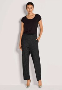 Vrouw staat in een zwart shirt met korte mouwen, gestructureerde zwarte high-waisted broek, zilveren hakken en hangende oorbellen, tegen een effen achtergrond.