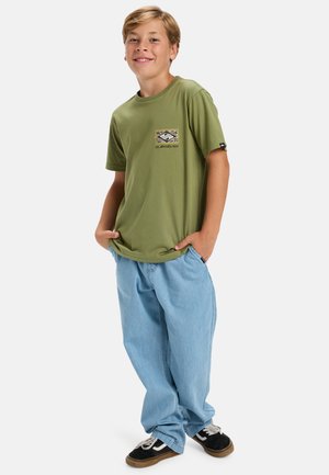 Olive green cotton t-shirt with a small patterned pocket design, paired with light blue baggy jeans and black sneakers.