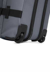 Valise à roulettes grise avec un tissu durable, des accents en plastique noir et des roues noires lisses. Les caractéristiques incluent des coins renforcés et des sangles robustes.