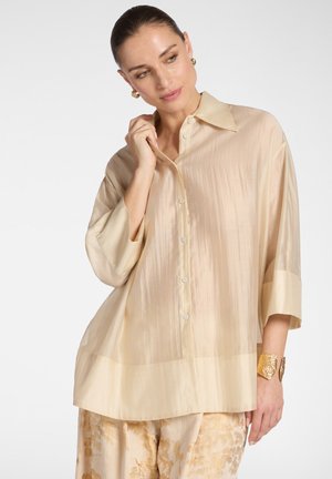 Vrouw draagt een losse beige satijnen blouse met wijde manchetten en beige broek met bloemenpatroon, gecombineerd met een gouden armband en oorringen.