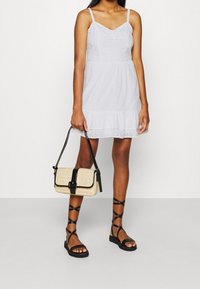 Vit, ärmlös klänning med texturerad tyg och volangkant, kombinerad med en liten, stråfärgad handväska med en svart rem och spänne.