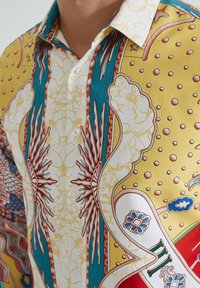 Chemise avec des motifs abstraits jaunes, blancs, bleus et rouges présentant des formes étoilées, des motifs floraux et des détails en points, boutonnée au col.