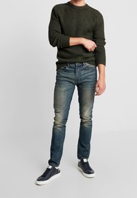 Ciemnozielony sweter z prążkowanej dzianiny, niebieskie sprane dżinsy i granatowe sneakersy. Slim-fit, okrągły dekolt i raglanowe długie rękawy.