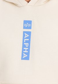 Felpa color beige con un logo rettangolare blu che presenta la parola "ALPHA" e un simbolo a stella, realizzata in un tessuto morbido e strutturato.
